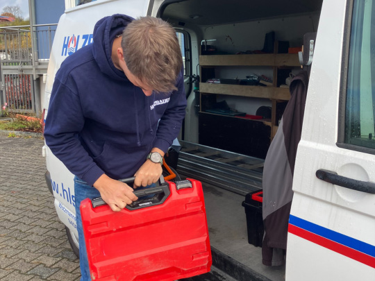 Der Geschäftsführer von der Firma Holzbauer steht vor seinem Transporter mit einem Werkzeugkoffer in der Hand