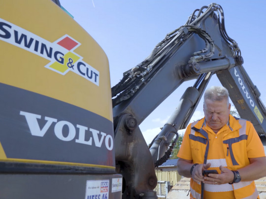 Mitarbeiter von SWING & CUT steht mit Smartphone in der Hand neben Bagger auf der Baustelle.